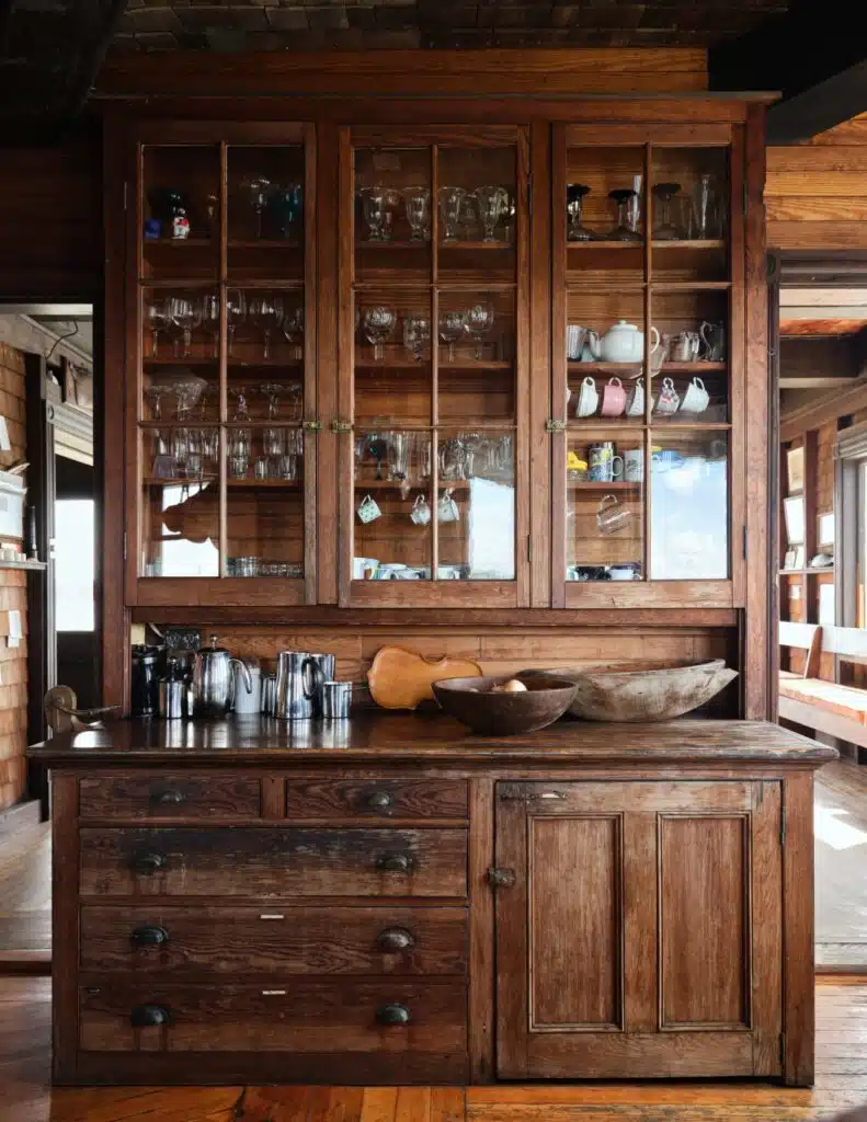 A rustic wooden hutch with glass cabinets holding dishes, glasses, bowls, pitchers, and cups.