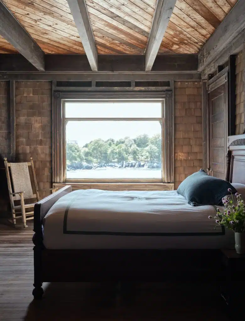 Rustic bedroom with wooden ceiling, large window with a water view, bed, rocking chair, and flowers.
