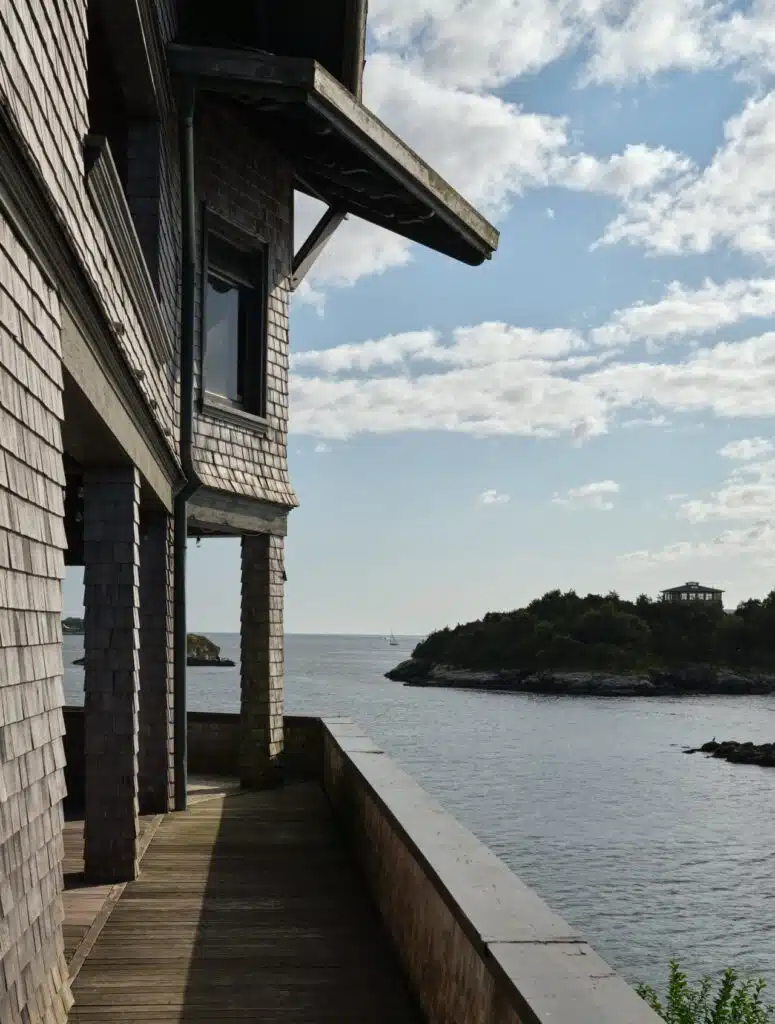Shingle-sided house with a wooden deck overlooks calm water and a tree-covered island under a partly cloudy sky.