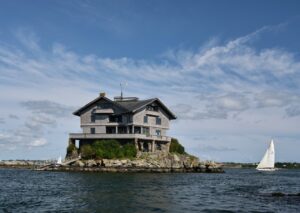 Clingstone. A large house sits on a rocky island with a sailboat passing nearby under a blue sky with clouds.