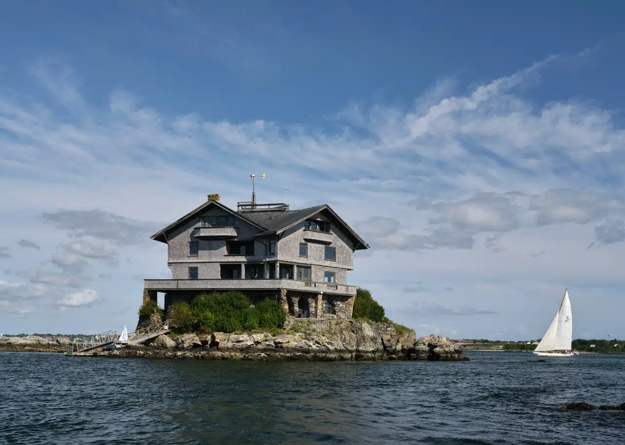 Clingstone. A large house sits on a rocky island with a sailboat passing nearby under a blue sky with clouds.