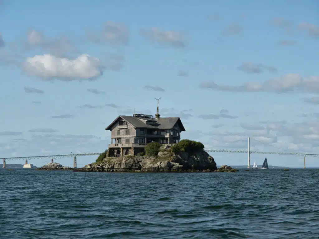 House on a small rocky island, surrounded by water, with a bridge and sailboats in the background.