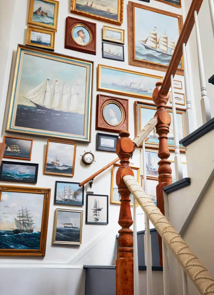 A staircase wall filled with framed paintings of ships and sailboats in various sizes and styles.