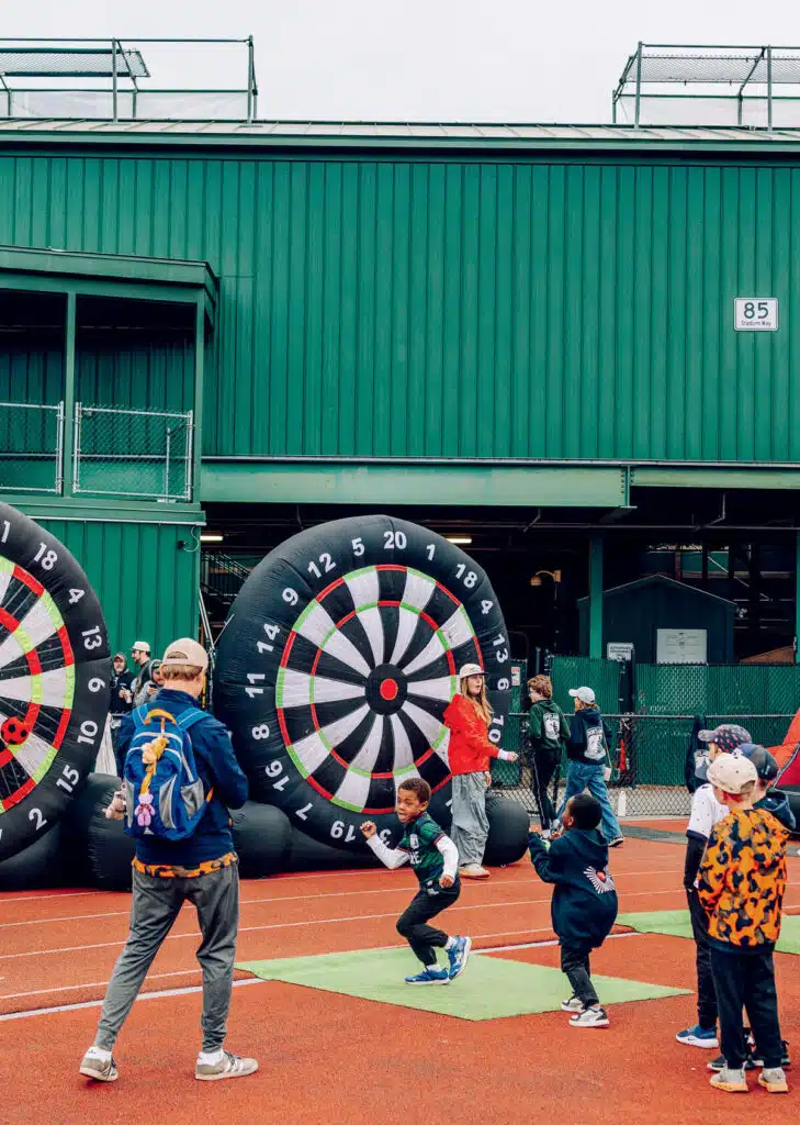 Children play a soccer dart game outdoors, aiming balls at a large inflatable dartboard.