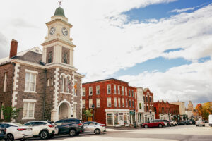 A historic clock tower with cars parked in front and brick buildings lining a sunny street.
