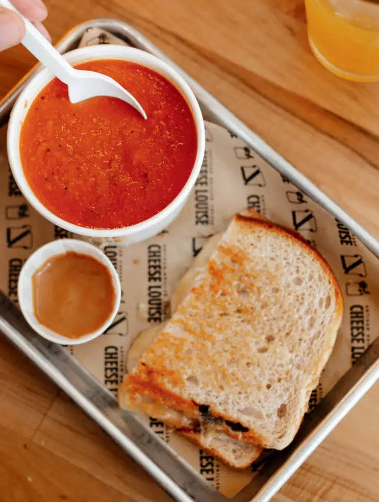 Grilled cheese sandwich on a tray with a bowl of tomato soup and a small cup of sauce.
