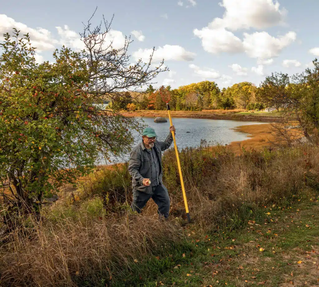 Person in outdoor clothes holding a long yellow pole near a pond, surrounded by autumn trees and grass.