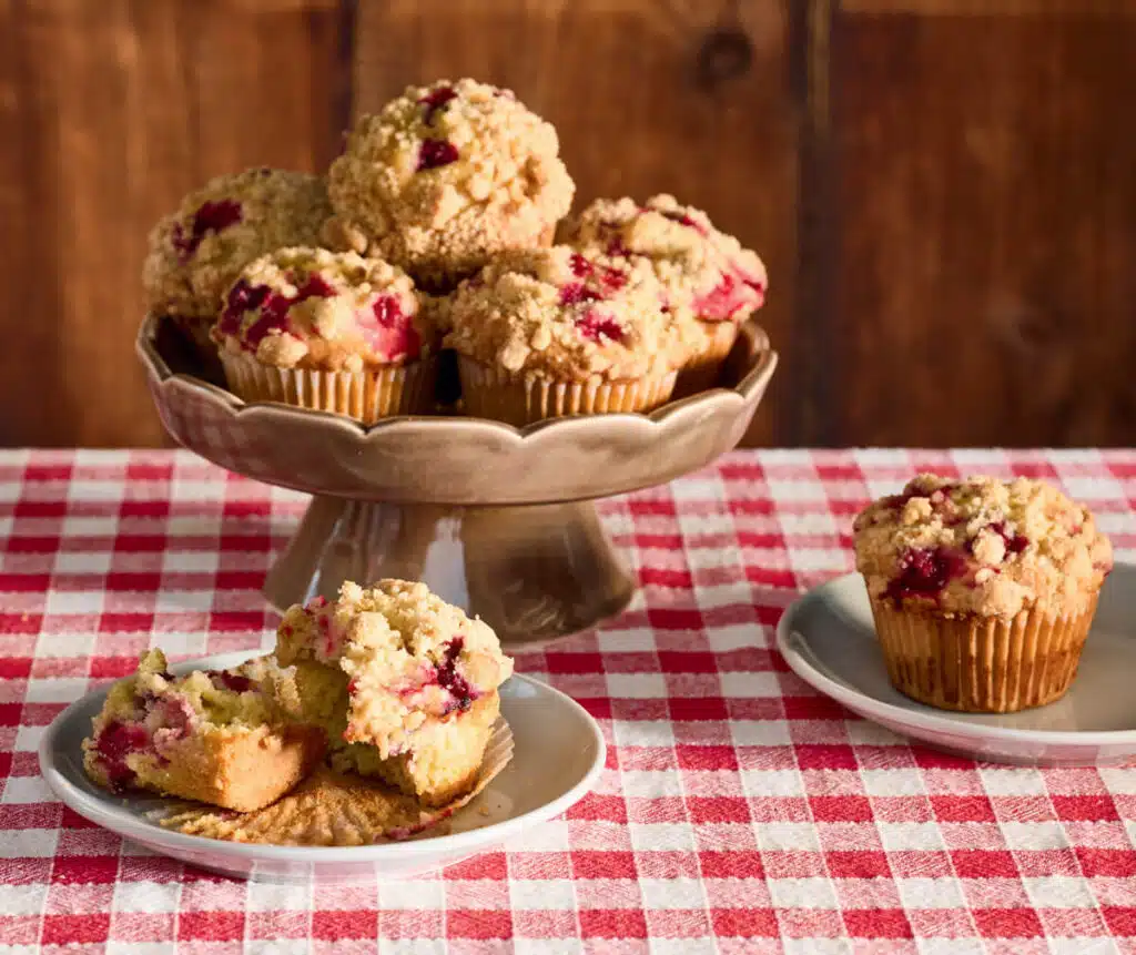 Cranberry-Orange Muffins with Streusel Topping. Cranberry muffins on a stand and plate, with one muffin cut open, on a red checkered tablecloth.