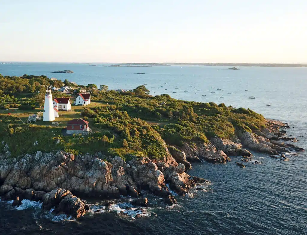 A lighthouse and houses sit on a rocky, green coastline overlooking the ocean under a clear sky.