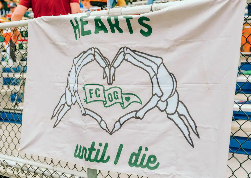 Inside the Rise of Portland’s Hearts of Pine Soccer Club. Banner with skeleton hands forming a heart, text reads “HEARTS until I die” and “FC OG” inside the heart.