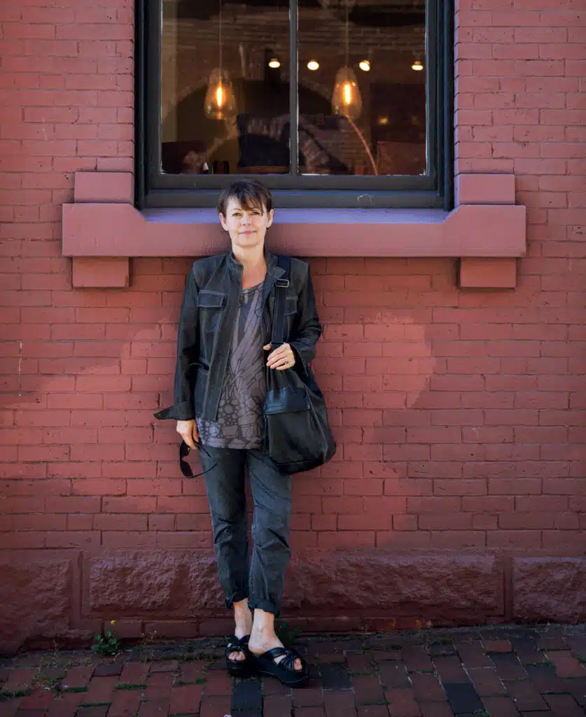 Made in New England: Rough & Tumble Bags in Portland, Maine. Woman in dark casual clothes stands against a red brick wall, holding a black bag and sunglasses.