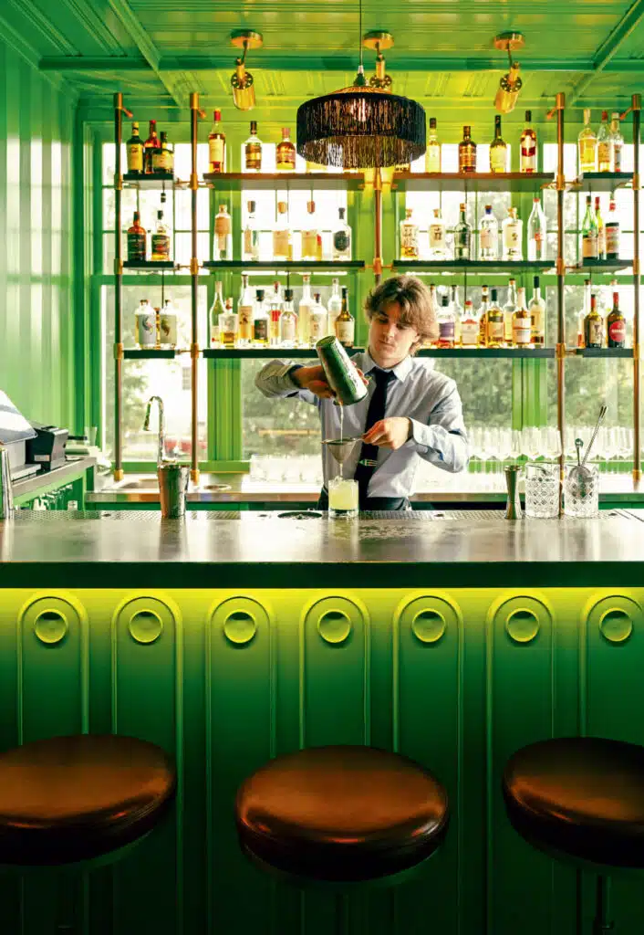 Bartender in a green bar pours a drink behind the counter, bottles and glasses on shelves behind.