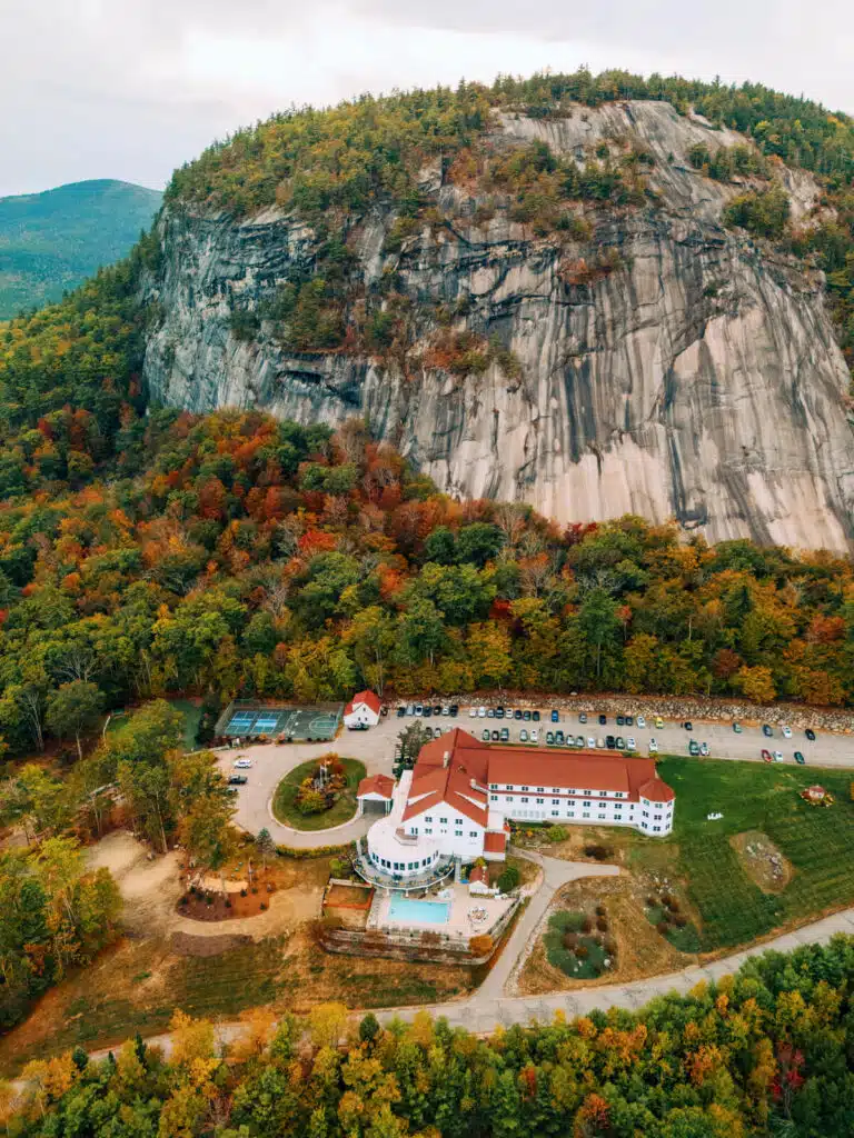 Fall Weekend in North Conway, New Hampshire. A white hotel with a red roof sits below a large rocky hill, surrounded by autumn trees.