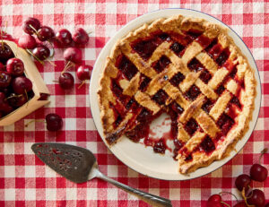 Triple Cherry Lattice Pie. Cherry pie with a lattice crust, partially eaten, on a red checkered tablecloth with fresh cherries.