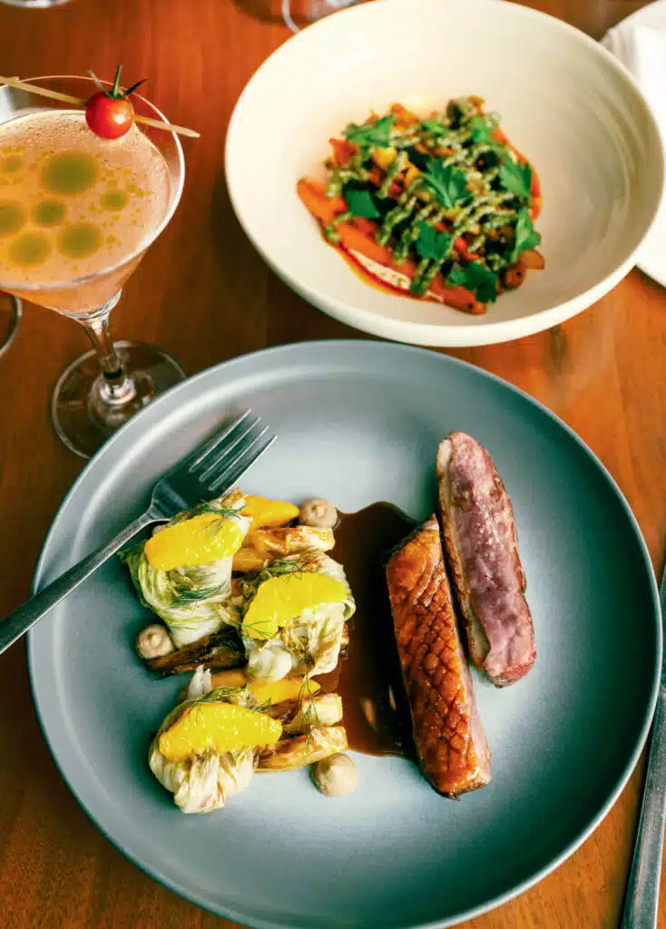 Plated duck breast with sauce, greens, and cocktail on a wooden table; salad dish in background.