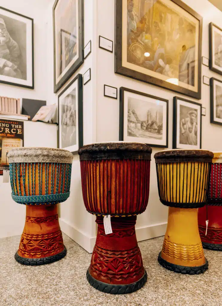 Three colorful African drums sit in an art gallery, surrounded by framed black-and-white artwork.