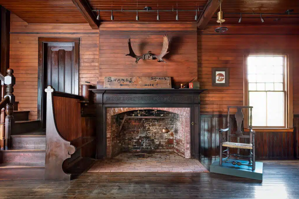 Rustic wooden room with brick fireplace, mounted antlers, rocking chair, and staircase on the left.