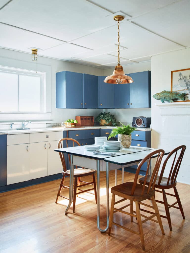 Bright kitchen with blue cabinets, a small dining table, wooden chairs, and sunlight through a window.