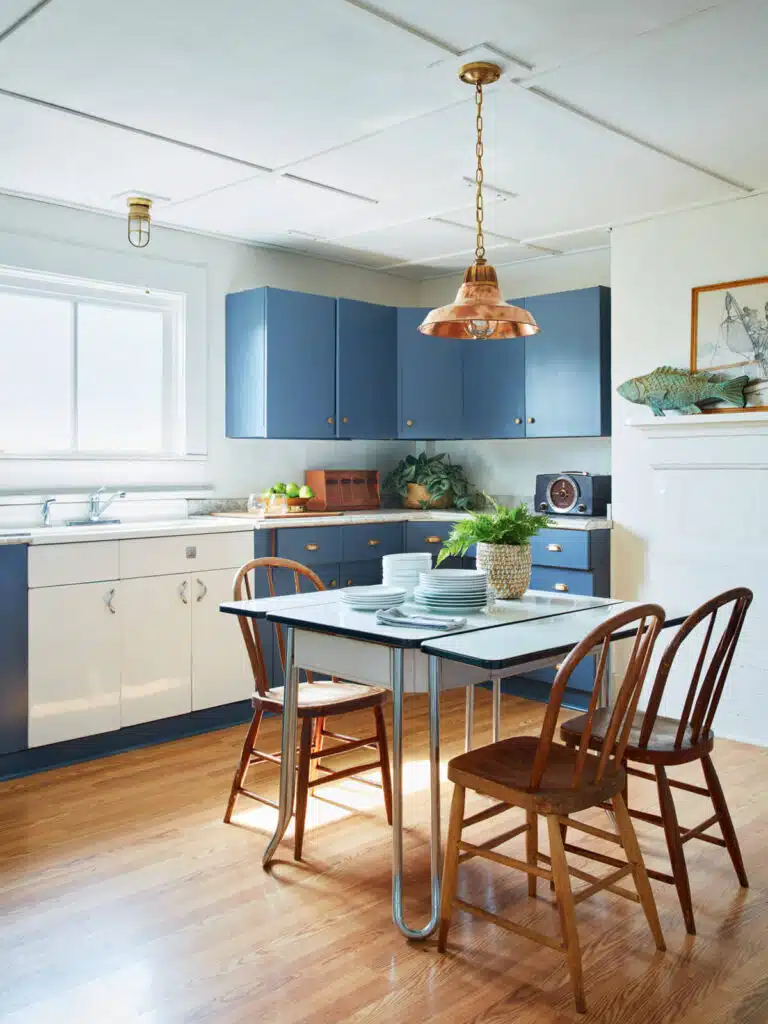 Bright kitchen with blue cabinets, a small dining table, wooden chairs, and sunlight through a window.