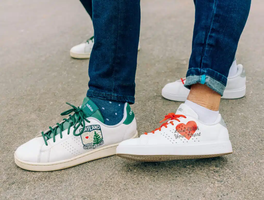 Two people wearing white Adidas sneakers with unique designs standing on gray pavement.
