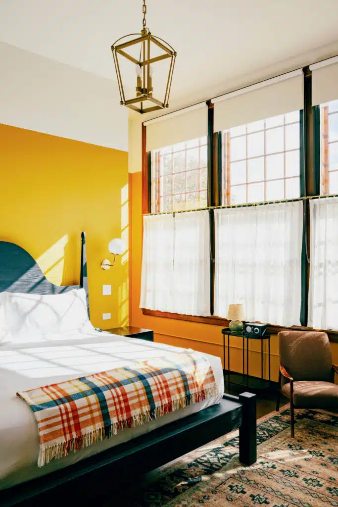 Sunny bedroom with yellow walls, large windows, a bed with plaid blanket, chair, and rug.
