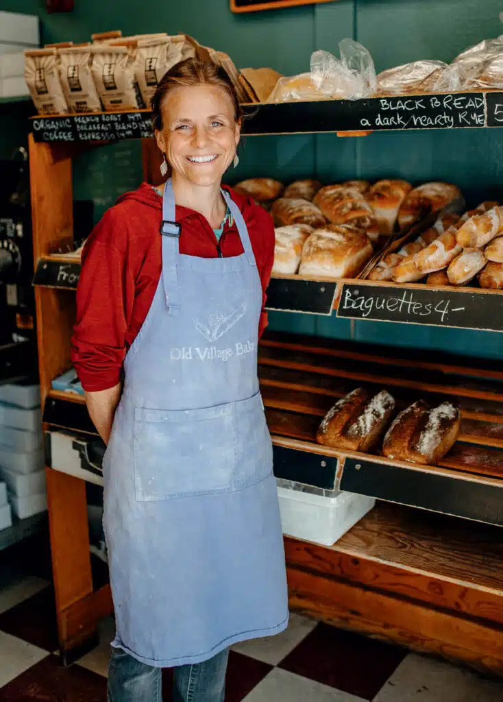 Smiling woman in a blue apron stands in front of shelves filled with bread at a bakery.