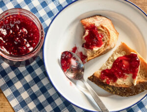 Raspberry Jam. Two bread slices with butter and raspberry jam on a plate, a spoon, and a jar of jam on a checkered cloth.