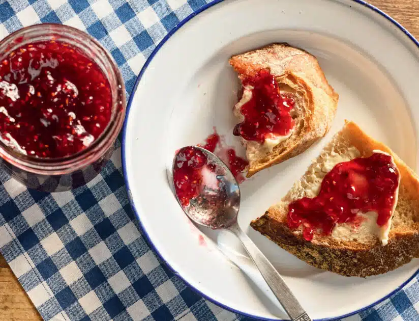 Raspberry Jam. Two bread slices with butter and raspberry jam on a plate, a spoon, and a jar of jam on a checkered cloth.