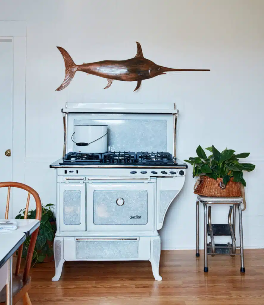 Vintage white stove with a pot on top, a fish sculpture above, and plants nearby on wooden floor.