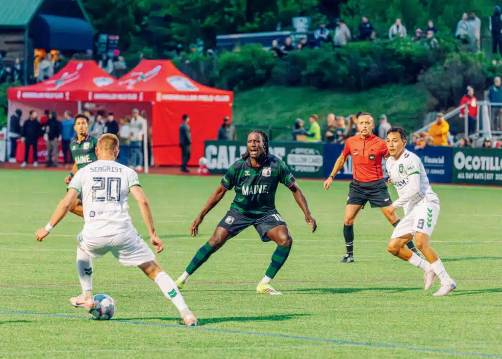 Soccer players compete for the ball on a field as a referee and spectators watch in the background.