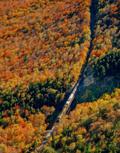 Fall Weekend in North Conway, New Hampshire. A train travels through a forested valley with vibrant autumn foliage in shades of orange, yellow, and green.