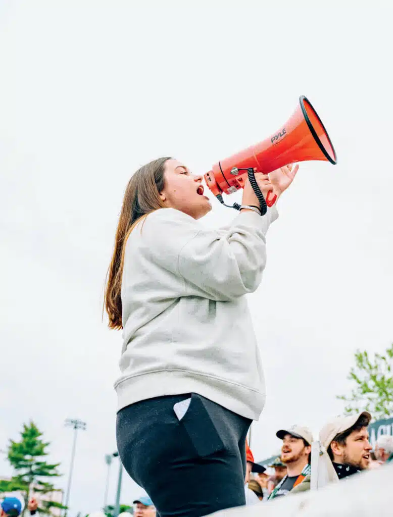 Person in a white sweatshirt shouting into a red megaphone at an outdoor event, with people around.