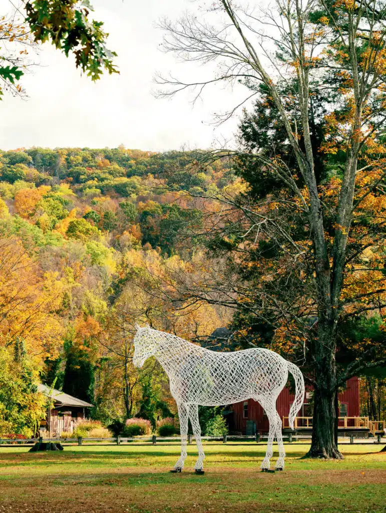 A large white wire horse sculpture stands on grass with autumn trees and a barn in the background.
