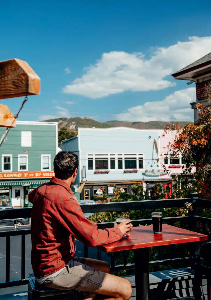 Man in a red shirt sits at a table on a balcony, holding a drink, overlooking colorful buildings.