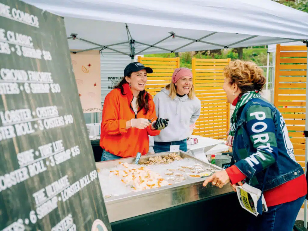Two vendors and a customer laugh together at an outdoor seafood stand with oysters and shrimp on ice.