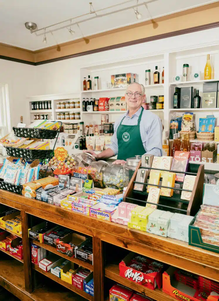 Smiling shopkeeper in apron stands behind a counter filled with candy and snacks in a quaint store.