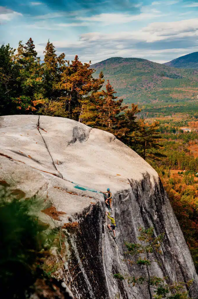 Fall Weekend in North Conway, New Hampshire. A rock climber ascends a steep granite cliff with trees and mountains in the background under a blue sky.