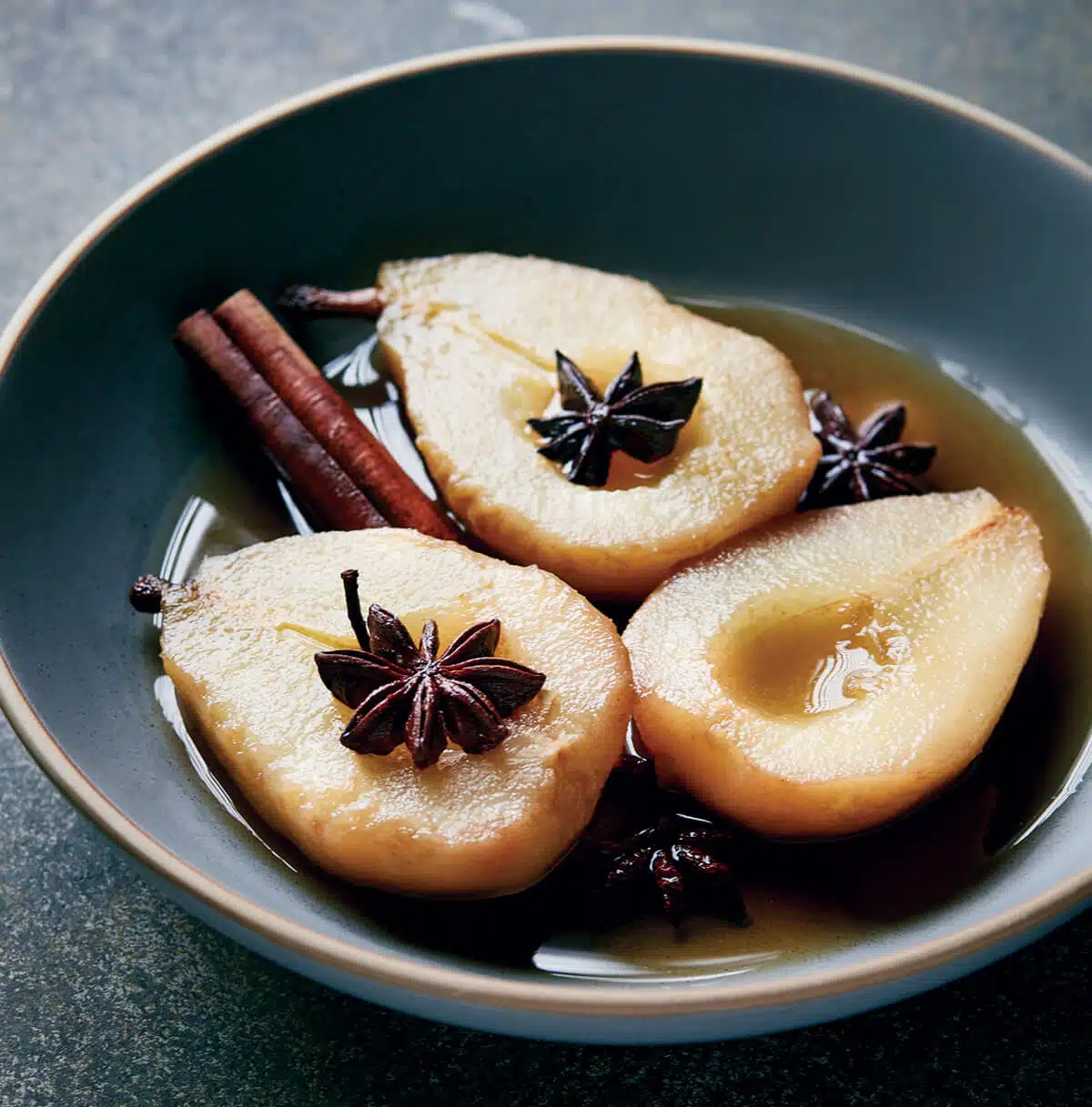 Halved poached pears in syrup with star anise and cinnamon sticks in a dark bowl.