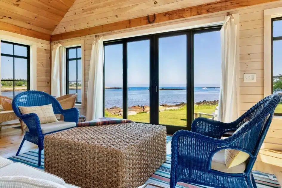Maine vacation rental's sunlit room with wicker and blue chairs, large windows, and ocean view.