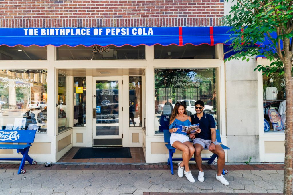 A couple sits on a bench reading outside the Birthplace of Pepsi Cola store on a sunny day.