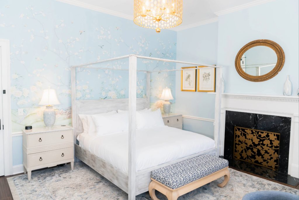 Elegant bedroom at The Harvey in New Bern with a canopy bed, blue floral wallpaper, fireplace, and two nightstands with lamps.