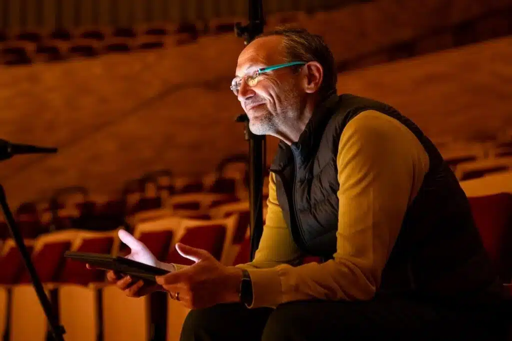 Man smiling while sitting in theater seats, holding a tablet with warm light illuminating his face.