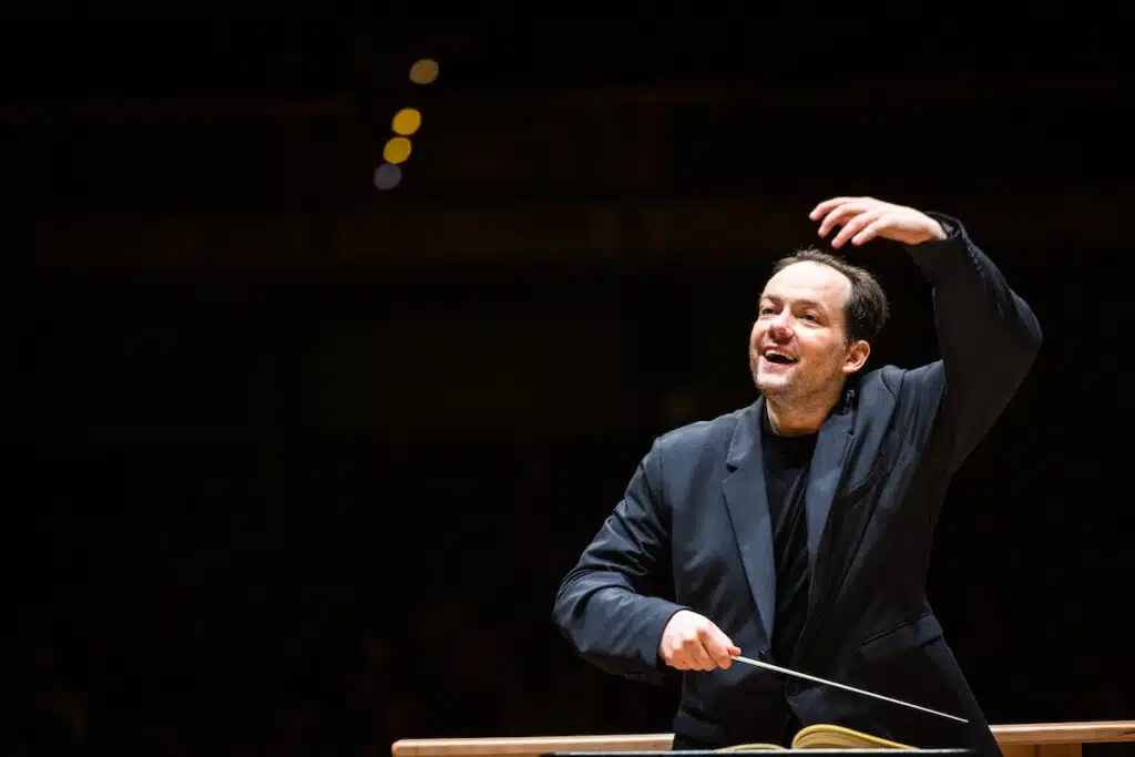 A conductor in a dark suit leads an orchestra, raising one hand and holding a baton in the other.