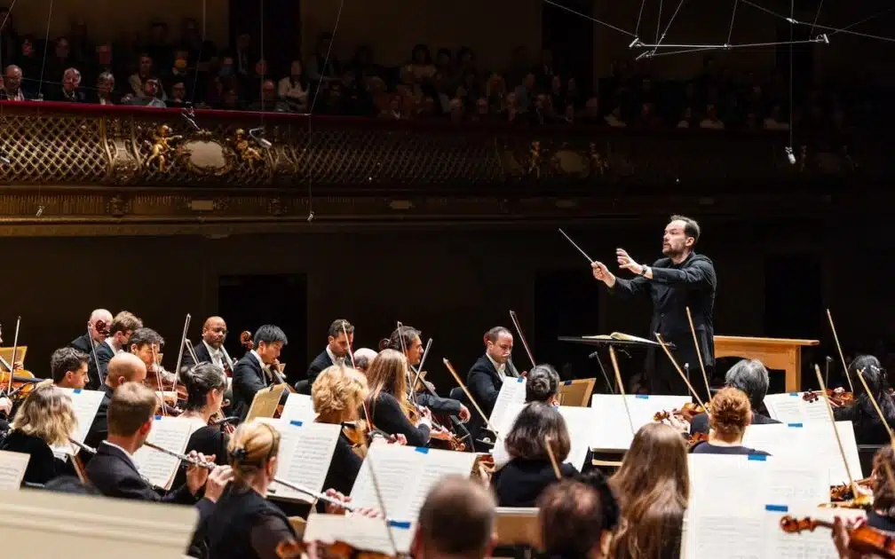 A conductor leads an orchestra during a live performance in a grand concert hall filled with an audience.