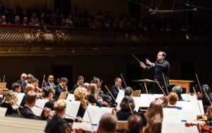 A conductor leads an orchestra during a live performance in a grand concert hall filled with an audience.