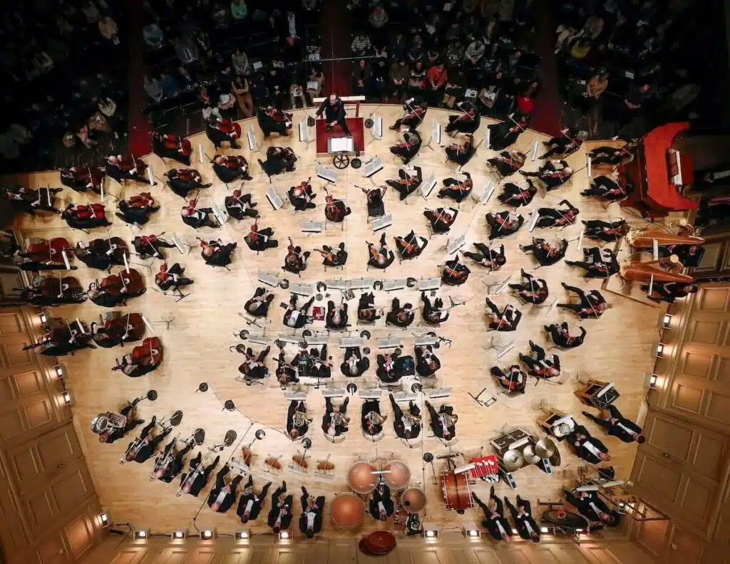 A symphony orchestra performs on stage, viewed from above, with musicians arranged in a semicircle.