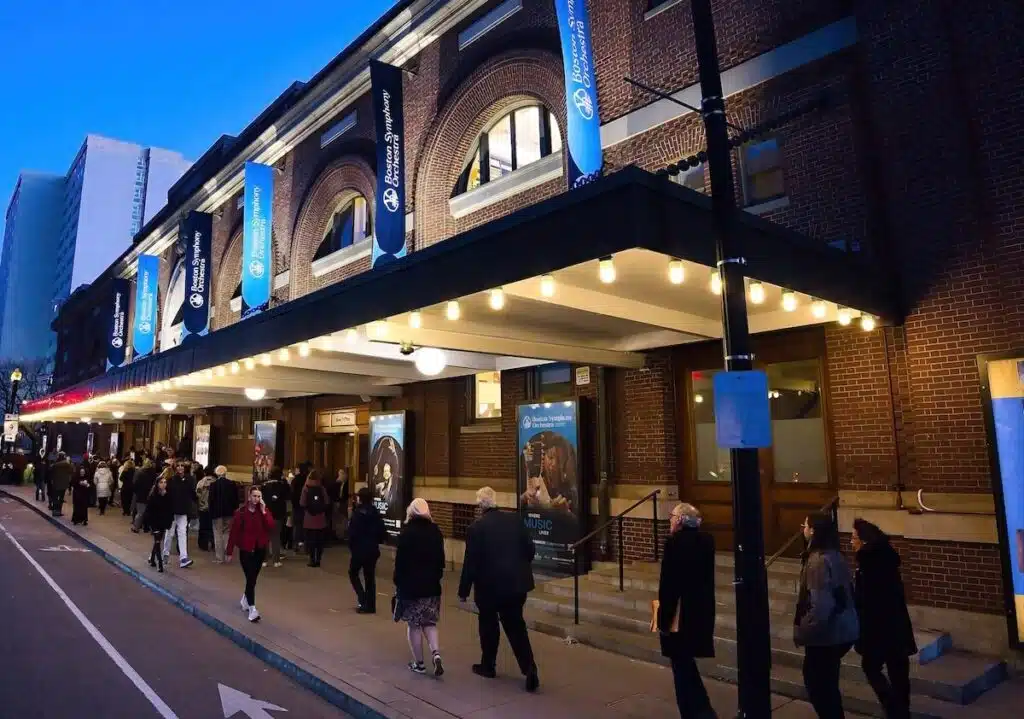 People walk into a theater with blue banners and lit marquee on a city street at dusk.