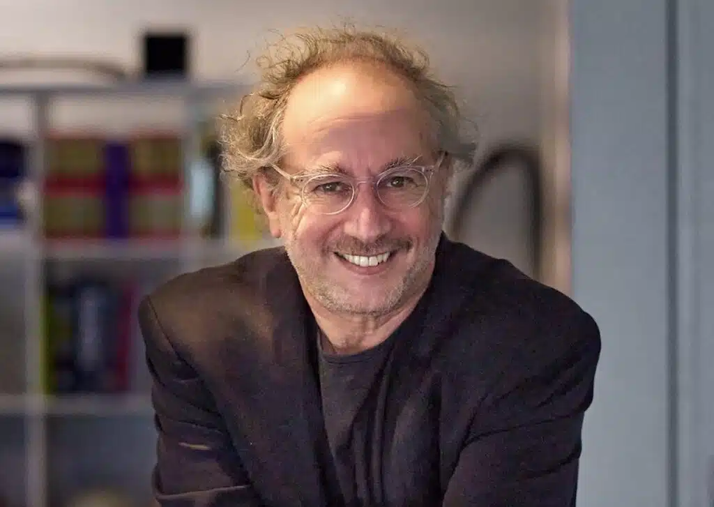 An older man with glasses and a black blazer smiles indoors, with shelves and books blurred in the background.