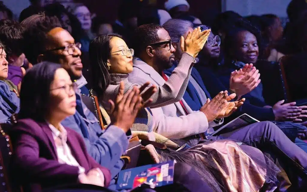Audience members seated closely together, clapping and smiling during an indoor event or performance.