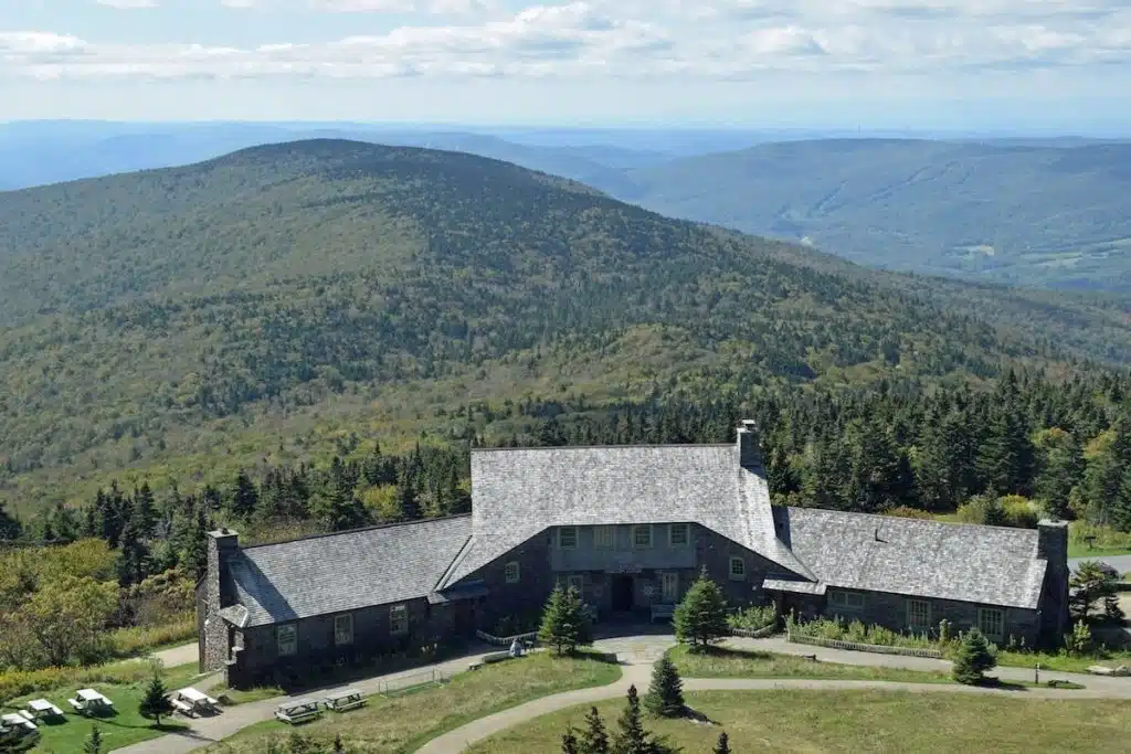 Stone lodge with gray roof on a green mountain, surrounded by trees and distant rolling hills.